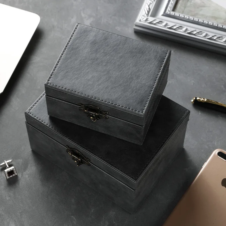 Two closed black signal blocking Faraday boxes for car keys to prevent keyless car theft stacked on a gray surface with a phone and photo frame in the background.